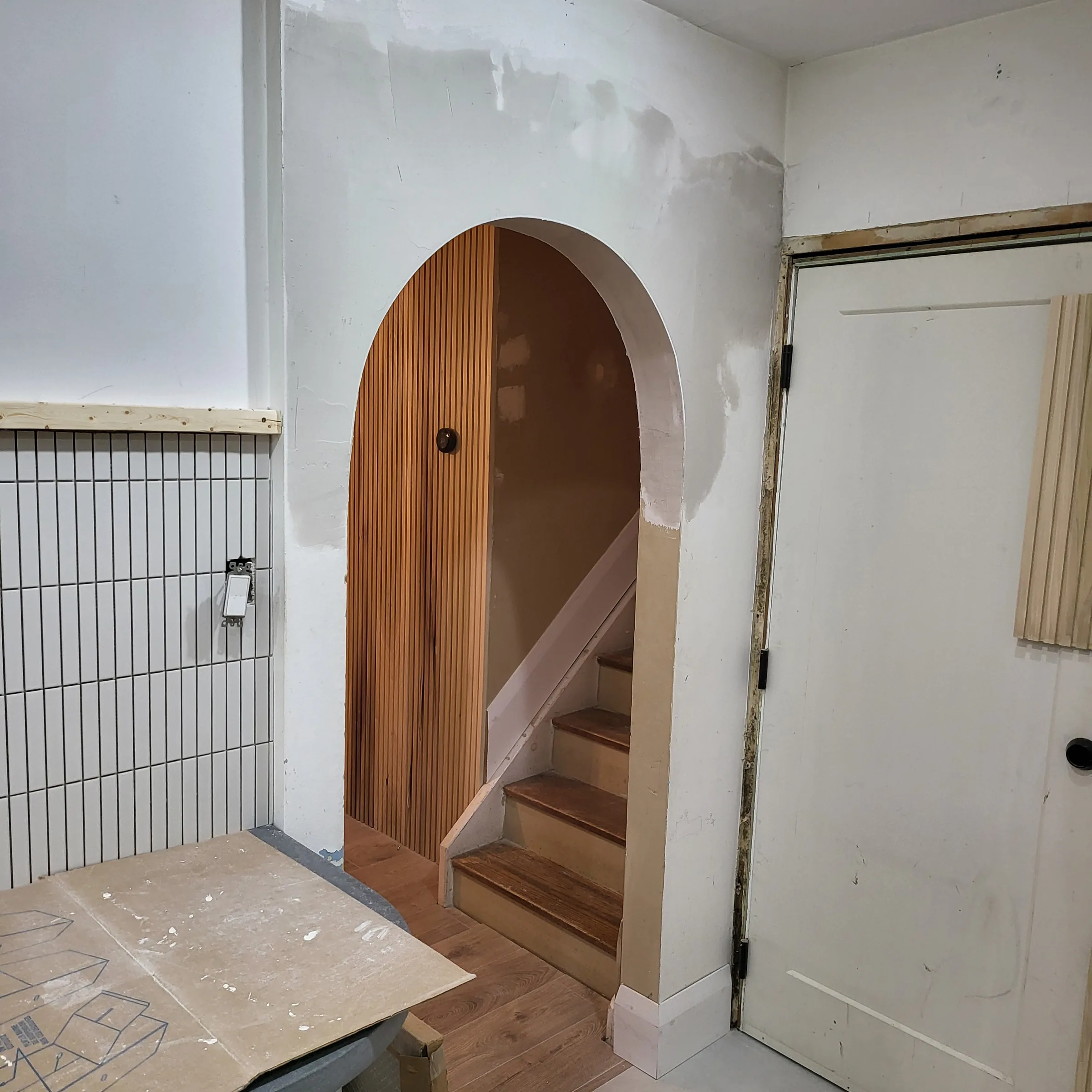 Framed and drywalled arched opening onto a staircase, with a vertical wood slat feature wall behind it