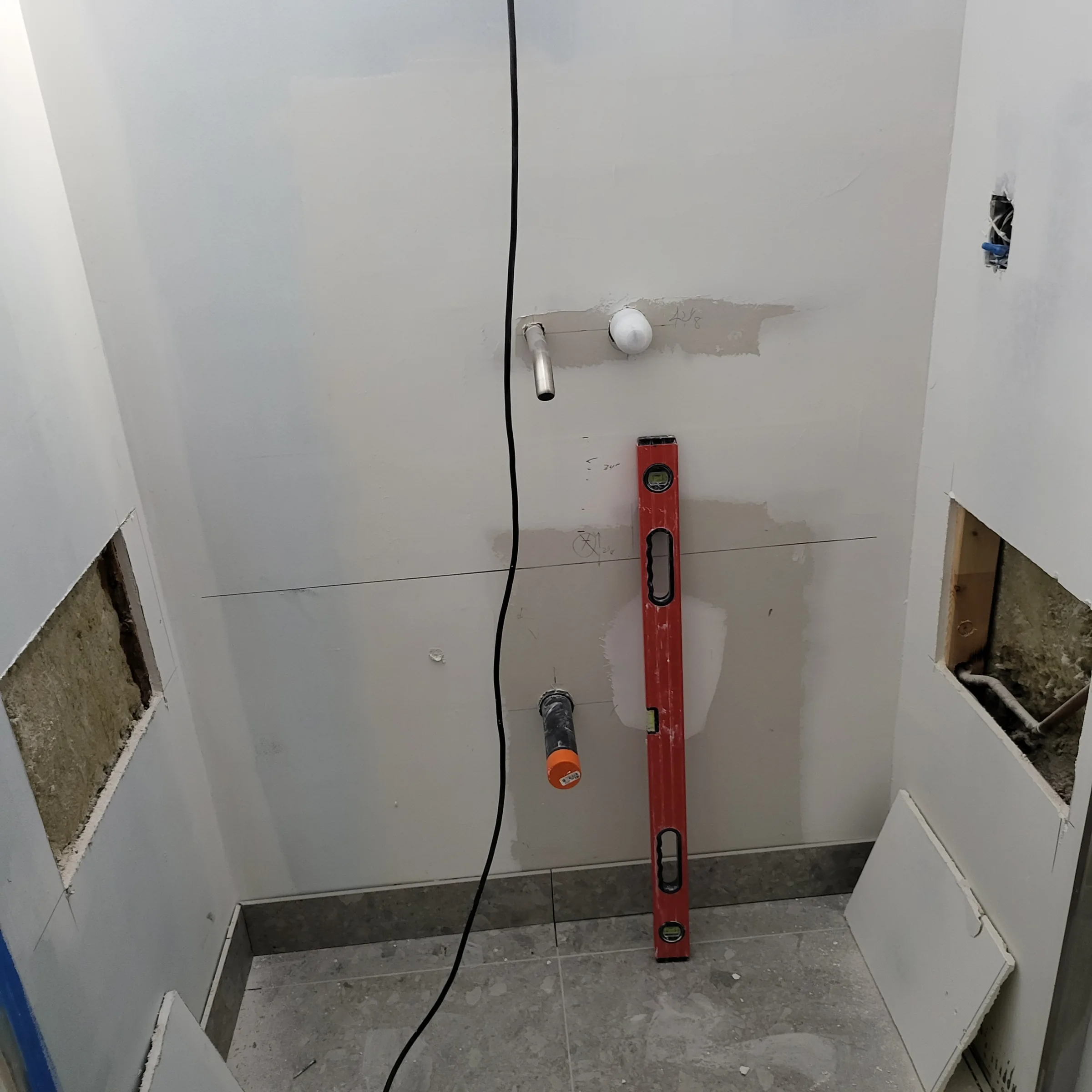 Bathroom wall at rough-in stage with drywall up, plumbing stubbed out and a level checking the layout