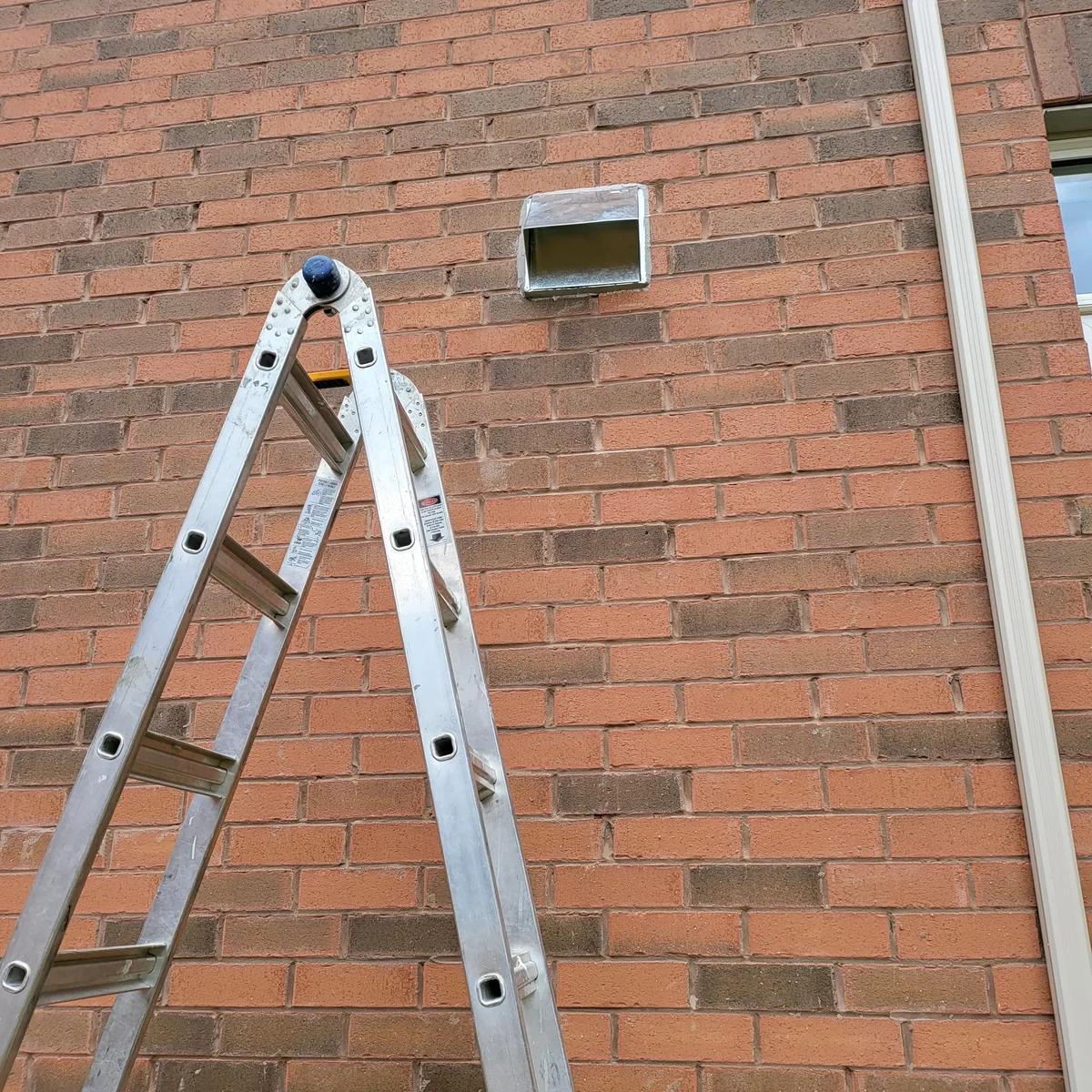 New exterior vent cap installed through a brick wall, with a ladder set up alongside